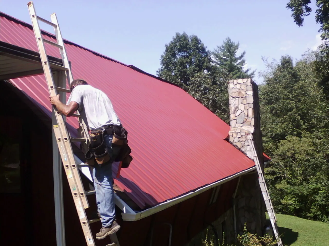 Roofer on a ladder beside a red metal roof and stone chimney — Soffit & Fascia Repair by Roofing Brothers in Manistee